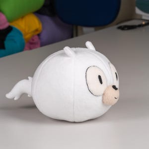 May include: White plush toy shaped like a round creature with a long tail. The toy has black eyes and a small, beige nose.