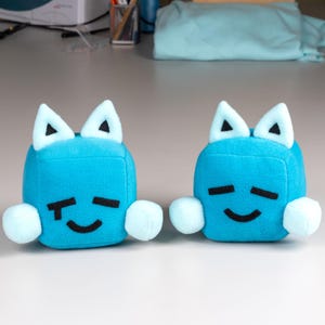 May include: Two light blue cube-shaped plush toys with white triangular ears and light blue pom-poms. Each toy has black eyes and a smiling mouth. The toys are designed to look like a cat.