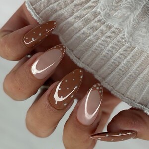 May include: Almond-shaped nails featuring a combination of solid brown polish with small white polka dots and French-style tips with a nude base and brown tips accented by white dots. The manicure displays a glossy finish on all fingernails.