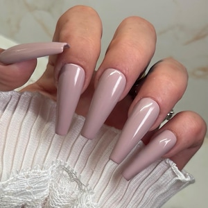 May include: Close-up of a hand with long, nude-colored, coffin-shaped acrylic nails. The nails are smooth and shiny, with a subtle shimmer.