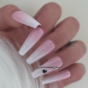 May include: Long, pink and white ombre press-on nails with a black heart and rhinestones on one nail.