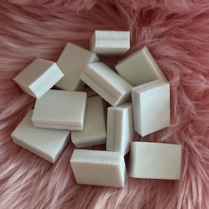 Könnte beinhalten: Ein Stapel weißer Nagelpolierblöcke auf einer rosa flauschigen Oberfläche. Die Blöcke sind rechteckig und haben auf einer Seite eine glatte Oberfläche und auf der anderen Seite eine raue Oberfläche.