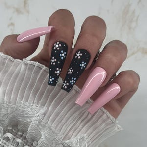 Könnte beinhalten: Eine Hand, die lange, Sarg-förmige Kunstnägel zeigt. Einige Nägel sind einfarbig hellrosa lackiert, während andere matt schwarz mit weißen, hellblauen und rosa floralen Designs sind. Die Nägel sind ein modisches Accessoire.
