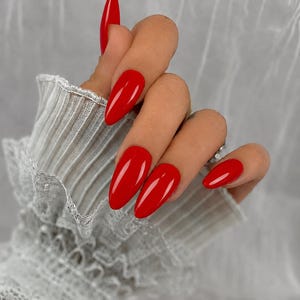 May include: Long, almond-shaped fingernails painted in a glossy, vibrant red polish. The nails feature a smooth, reflective finish and are displayed against a white, ribbed, long-sleeved garment with a ruffled, lace-trimmed cuff detail.