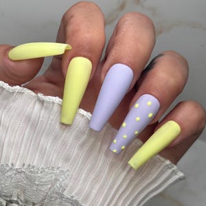 Könnte beinhalten: Eine Hand zeigt lange, matte, Sarg-förmige Kunstnägel. Die Nägel sind in Pastellgelb und Lavendel lackiert. Ein lila Nagel hat gelbe Tupfen. Die Nägel sind ein Modeaccessoire.