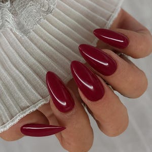 Könnte beinhalten: Lange, mandelförmige Fingernägel mit einem glänzenden, tief burgunderroten Nagellack-Finish. Die Nägel befinden sich in der Nähe einer weißen, plissierten, halbtransparenten Stoffmanschette mit zarter Spitzenborte am oberen Rand.