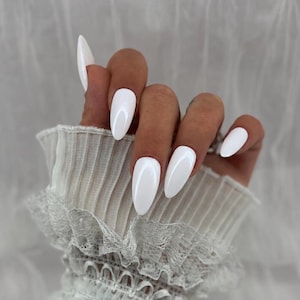 May include: A set of almond-shaped press-on nails with a glossy, solid white finish. The nails are displayed on fingers, showcasing a smooth, reflective surface and a pointed tip design, paired with a white pleated fabric sleeve with lace trim.