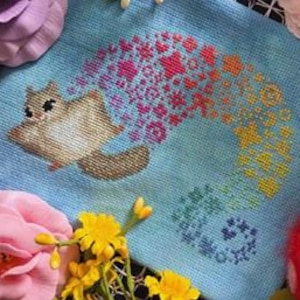 May include: A cross-stitch embroidery on light blue fabric featuring a brown flying squirrel with a trailing rainbow-colored pattern of stars and geometric shapes. The text "BAD Stitch FIBER ARTS + ZINES" and "Flying Squirrel" appear on the page.