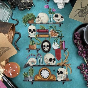 May include: A cross-stitch design featuring a shelf with skulls, plants, books, and a clock. The color palette includes white, green, brown, and black. The design has a gothic or witchy theme.