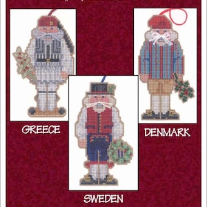 May include: Three nutcracker ornaments, each representing a different country. The first is a Greek nutcracker wearing a white and blue outfit with a red hat. The second is a Swedish nutcracker wearing a red vest, blue pants, and a black hat. The third is a Danish nutcracker wearing a blue and red striped shirt with a red hat. The ornaments are all part of a set called "Nutcracker Ornaments IV" designed by Sandra Cozzolino.
