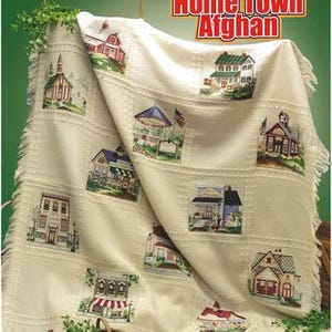 May include: A cream-colored afghan featuring embroidered squares with various buildings, including houses, a church, and a gazebo. The afghan has a fringed edge and is displayed with a basket of greenery. The text "Home Town Afghan" is visible.