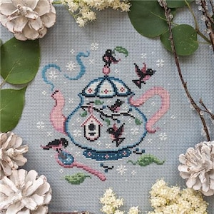 May include: Cross-stitch design featuring a teapot with birds, a birdhouse, and floral accents. The teapot is light blue with pink and dark blue details. The design includes a pink handle and spout, with a decorative border and a bird perched on top.