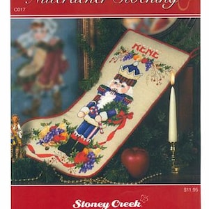 May include: A counted cross stitch pattern for a Christmas stocking. The design features a nutcracker soldier in blue and red, surrounded by grapes and berries. The stocking is labeled "Nutcracker Stocking" and includes the text "Stoney Creek Collection."