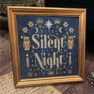 May include: A framed cross-stitch design with a blue background and white lettering that reads "Silent Night". The design features two owls wearing Santa hats, candles, stars, and crowns.