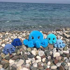 May include: Four crocheted octopus plushies in shades of blue, arranged on a bed of pebbles. The largest octopus is a bright blue with white eyes and a happy expression. The other three octopuses are smaller and have a more muted blue color.