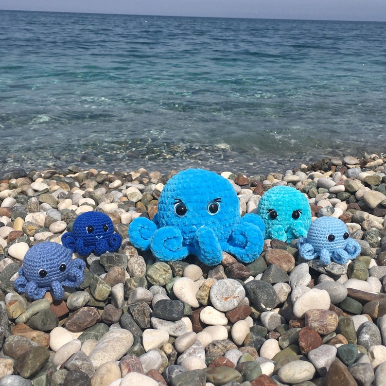 May include: Four crocheted octopus plushies in shades of blue, arranged on a bed of pebbles. The largest octopus is a bright blue with white eyes and a happy expression. The other three octopuses are smaller and have a more muted blue color.