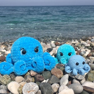 May include: Three crocheted octopus plushies in blue and light blue colors. The largest octopus is blue with white eyes and a light blue smile. The two smaller octopuses are light blue with white eyes. The plushies are sitting on a bed of gray and brown pebbles.