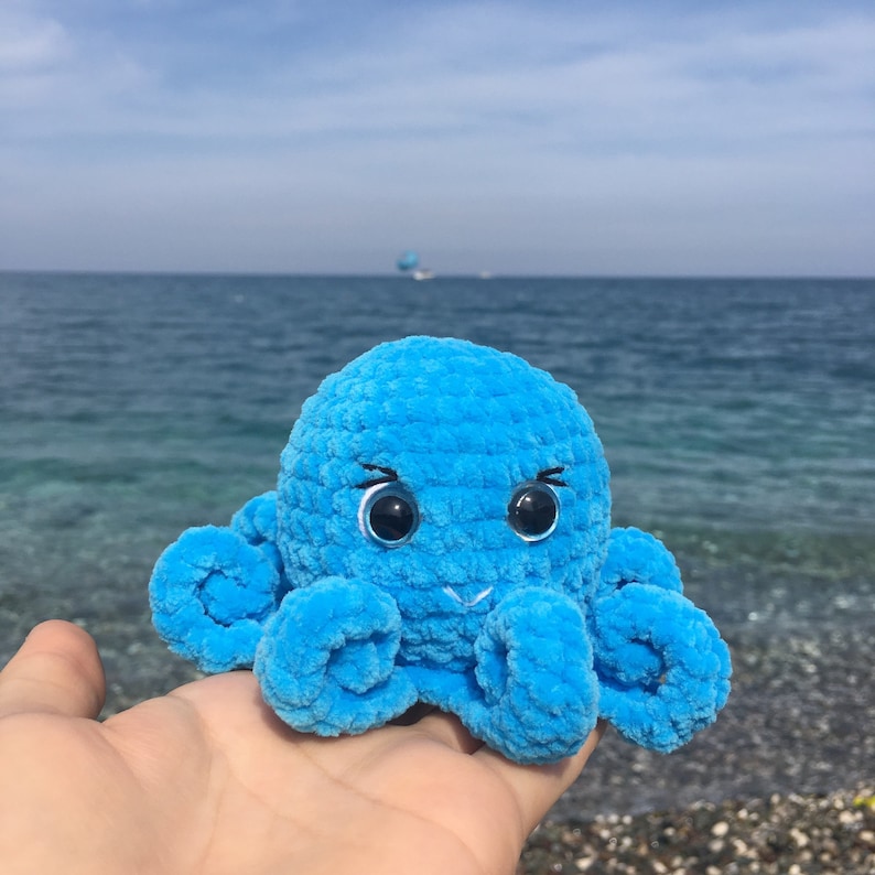 May include: A blue crocheted octopus with large black eyes. The octopus is sitting on a hand against a background of blue water.