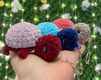 May include: Five crocheted turtle keychains in different colors: purple, blue, red, pink, and gray. The keychains are attached to a silver chain with a heart-shaped charm.