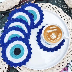 Könnte beinhalten: Eine Sammlung von gehäkelten Untersetzern in Blau, Weiß und Türkis, jeweils mit einem Augenmotiv. Auf einem Untersetzer steht eine weiße Tasse mit Kaffee und Latte Art. Die Untersetzer befinden sich auf einem weißen Teller und einem geflochtenen Platzset.