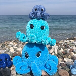 May include: Three crocheted octopus plushies stacked on top of each other. The top octopus is blue, the middle octopus is light blue, and the bottom octopus is a darker blue. The plushies are sitting on a bed of small, smooth, gray rocks.