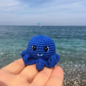 May include: A blue crocheted octopus with black eyes and a white smile. The octopus is sitting on a hand against a blurry background of blue water.