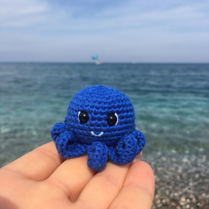 May include: A blue crocheted octopus with black eyes and a white smile. The octopus is sitting on a hand against a blurry background of blue water.