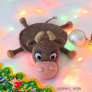 May include: A brown crocheted bull-shaped rug with a pink nose and white eyes. The rug is lying on a white surface with colorful lights in the background.