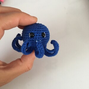 May include: A small blue crocheted octopus with black eyes. The octopus has eight tentacles and is being held by a hand.