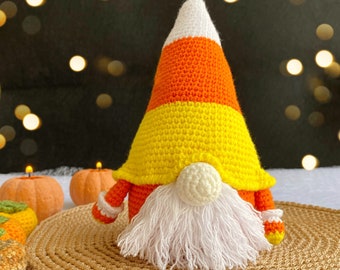 May include: A crocheted gnome wearing a candy corn hat. The gnome is sitting on a woven brown mat.