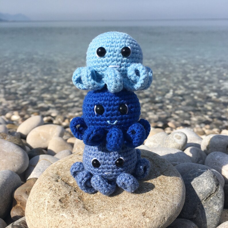 May include: A stack of three crocheted octopus plushies. The top octopus is light blue, the middle octopus is dark blue, and the bottom octopus is a lighter shade of blue. The octopuses are sitting on a gray rock.