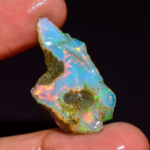 Op de afbeelding: Een ruwe opaal edelsteen met een levendig kleurenspel. De steen toont een mix van blauwe, oranje en groene tinten, met een melkachtige witte basis. De opaal wordt tussen vingers gehouden, wat de natuurlijke, onbewerkte vorm benadrukt.