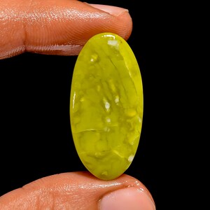 Pode incluir: Uma pedra preciosa oval polida, com um tom verde-amarelo vibrante. A pedra apresenta inclusões brancas e uma superfície brilhante. Segurada entre os dedos contra um fundo preto, destacando sua cor e textura.