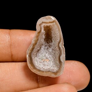 May include: A polished, open geode with a light brown and white exterior. The interior reveals a sparkling, crystalline cavity with shades of white and tan. The geode is held in a hand against a black background.