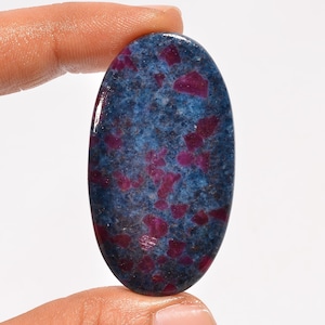 Op de afbeelding: Een ovaalvormige cabochon edelsteen met een diepblauwe basiskleur en verspreide, onregelmatige magenta en donkerrode insluitingen over het hele oppervlak. De steen heeft een gladde, gepolijste afwerking die het licht op het oppervlak weerkaatst.