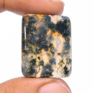 May include: A rectangular moss agate gemstone, held between fingers. The stone features a translucent base with intricate patterns of green, black, and yellow inclusions, resembling a miniature landscape. The polished surface reflects light, highlighting the natural variations.