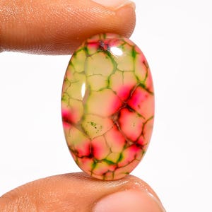 Puede incluir: Gema ovalada con un patrón llamativo. La piedra presenta una mezcla de colores rosa, verde y amarillo, con un diseño agrietado en forma de telaraña. La gema se sostiene entre los dedos, mostrando su superficie pulida y su combinación de colores única.