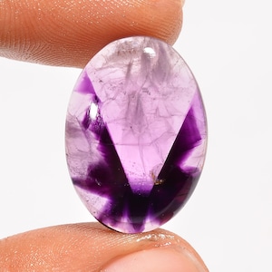 Może przedstawiać: Owalny kamień szlachetny ametystu o wypolerowanej powierzchni. Klejnot charakteryzuje się gradientem kolorów, przechodzącym od jasnego odcienia lawendy do głębokiego fioletu. Wewnętrzna struktura wykazuje unikalny, gwiaździsty wzór.