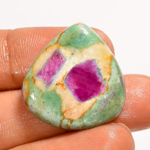 May include: A polished gemstone with a teardrop shape. The stone features a vibrant mix of colors, including a light green base with patches of cream and orange. Two prominent, deep pink ruby inclusions are visible within the stone.