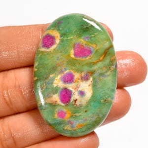 May include: An oval gemstone with a vivid green base, featuring bright pink and white inclusions. The polished surface of the stone reflects light, and the pink inclusions are of varying shapes and sizes.