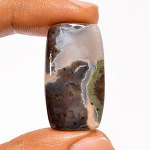 May include: A polished gemstone with a rectangular shape and rounded edges. The stone features a mix of colors, including white, gray, brown, and green, with a marbled appearance. The gemstone is held between two fingers.