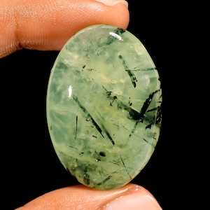 May include: An oval gemstone in a translucent, pale green colour. It has dark, needle-like inclusions creating a distinctive pattern. The polished surface reflects light, highlighting the internal structures. The gemstone is held between fingers against a black background.
