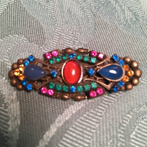 May include: A vintage brooch with a gold-tone metal setting and colourful stones. The brooch features three large stones: a red oval stone, a blue teardrop-shaped stone, and another blue teardrop-shaped stone. The brooch is decorated with smaller stones in shades of blue, green, and pink.