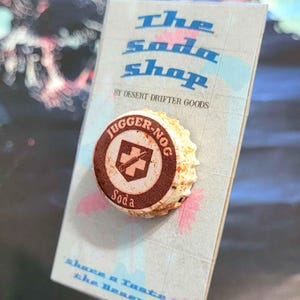 Könnte beinhalten: Ein Flaschenverschluss-Pin mit dem Text "JUGGER-NOG Soda" und einem Schild- und Kreuz-Design. Der Pin befindet sich auf einer Karte mit der Aufschrift "The Soda Shop" und "share a Taste of the Desert!"