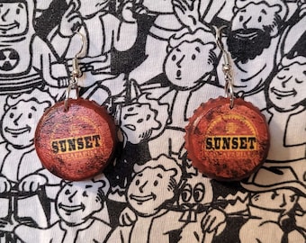 Może przedstawiać: Para ręcznie robionych kolczyków z kapsli z nadrukiem słowa "SUNSET". Kolczyki mają rustykalny wygląd z czerwoną i żółtą kolorystyką. Tło to czarno-biały nadruk graficzny.