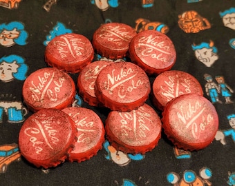 May include: A collection of red bottle caps with the words "Nuka Cola" printed in white. The bottle caps are arranged in a pile on a black fabric background with various patterns.