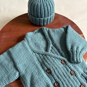 Könnte beinhalten: Ein tealfarbenes Strick-Babyset, bestehend aus einer gerippten Mütze und einer Strickjacke mit Holzknöpfen. Das Set wird auf einer Holzoberfläche präsentiert. Die Strickjacke hat lange Ärmel und einen V-Ausschnitt.