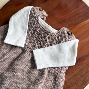 K&ouml;nnte beinhalten: Ein gestrickter Baby-Strampler in einem ged&auml;mpften Mauve-Ton, mit wei&szlig;em Kragen und Manschetten. Der Strampler hat ein strukturiertes Strickmuster und wird mit zwei kleinen, runden Kn&ouml;pfen befestigt. Das Kleidungsst&uuml;ck wird auf einer Holzoberfl&auml;che pr&auml;sentiert.