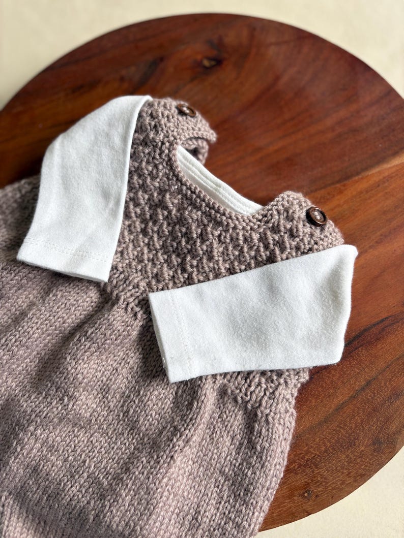 K&ouml;nnte beinhalten: Ein gestrickter Baby-Strampler in einem ged&auml;mpften Mauve-Ton, mit wei&szlig;em Kragen und Manschetten. Der Strampler hat ein strukturiertes Strickmuster und wird mit zwei kleinen, runden Kn&ouml;pfen befestigt. Das Kleidungsst&uuml;ck wird auf einer Holzoberfl&auml;che pr&auml;sentiert.