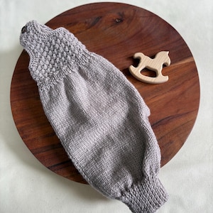 K&ouml;nnte beinhalten: Ein hellgrauer gestrickter Babysleeve mit strukturiertem Joch und Knopfverschluss. Ein kleines Holzschaukelpferd-Spielzeug befindet sich neben dem Sleeve. Der Sleeve liegt auf einem dunkelbraunen Holzbrett.